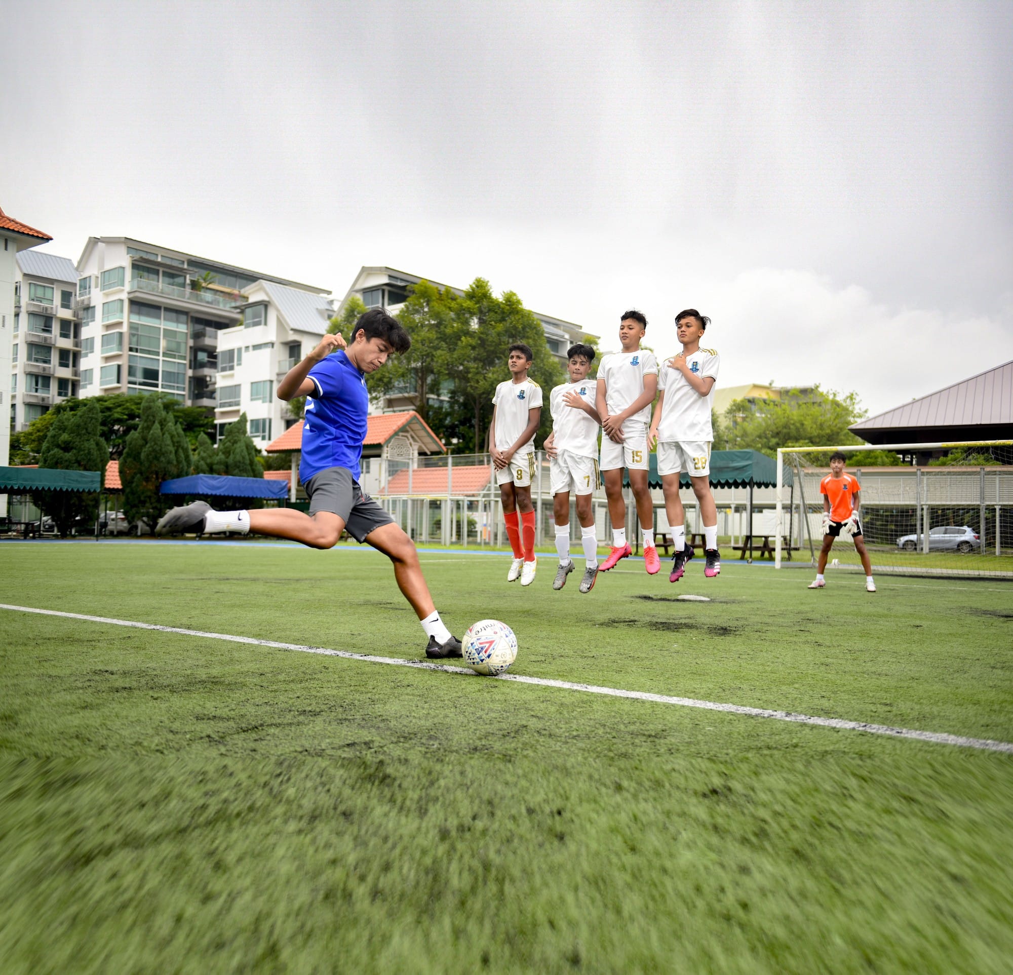 A boy in blue kicks a soccer ball as four players in white jump to form a wall on a green field.
